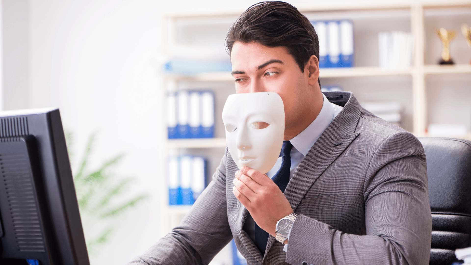 Photo of a businessperson at their desk holding a mask near their face as they look at their computer.