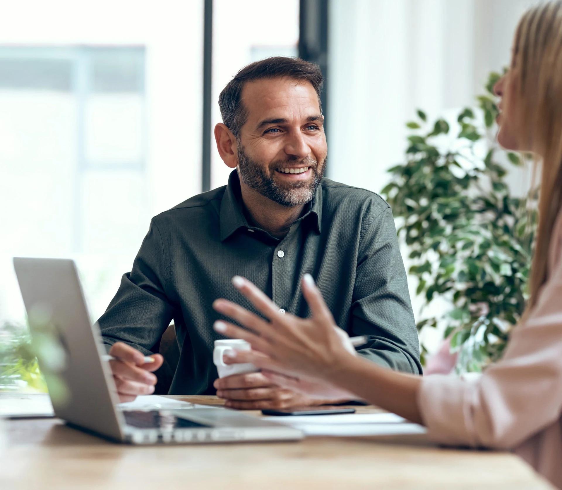 Photo of a person talking to another person in front of a laptop at an office.