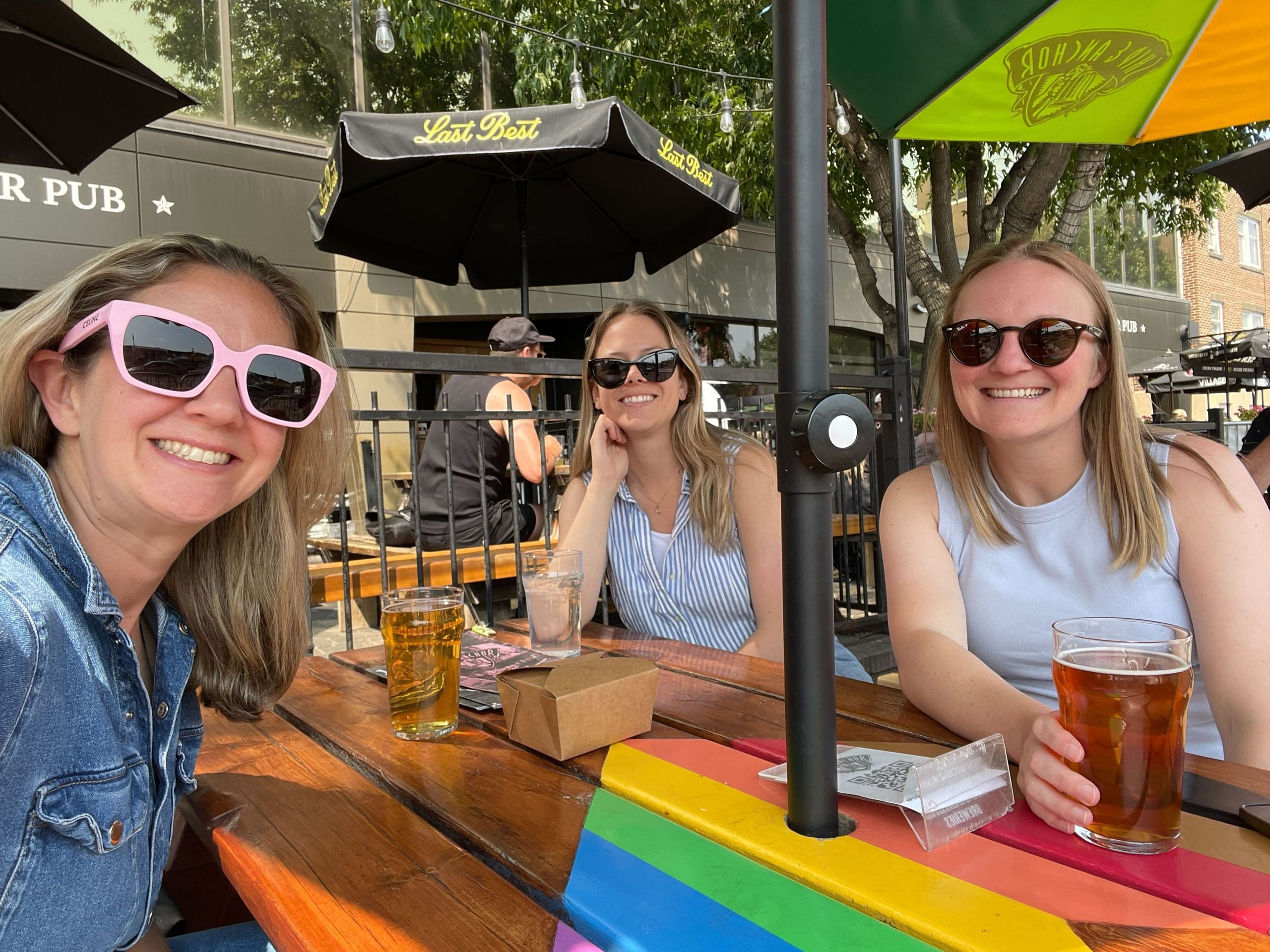 Three women smile and enjoy drinks at an outdoor patio on a sunny day. They are seated at a wooden picnic table painted with rainbow stripes, under umbrellas. Each woman is wearing sunglasses and appears relaxed and cheerful, with beverages in front of them.
