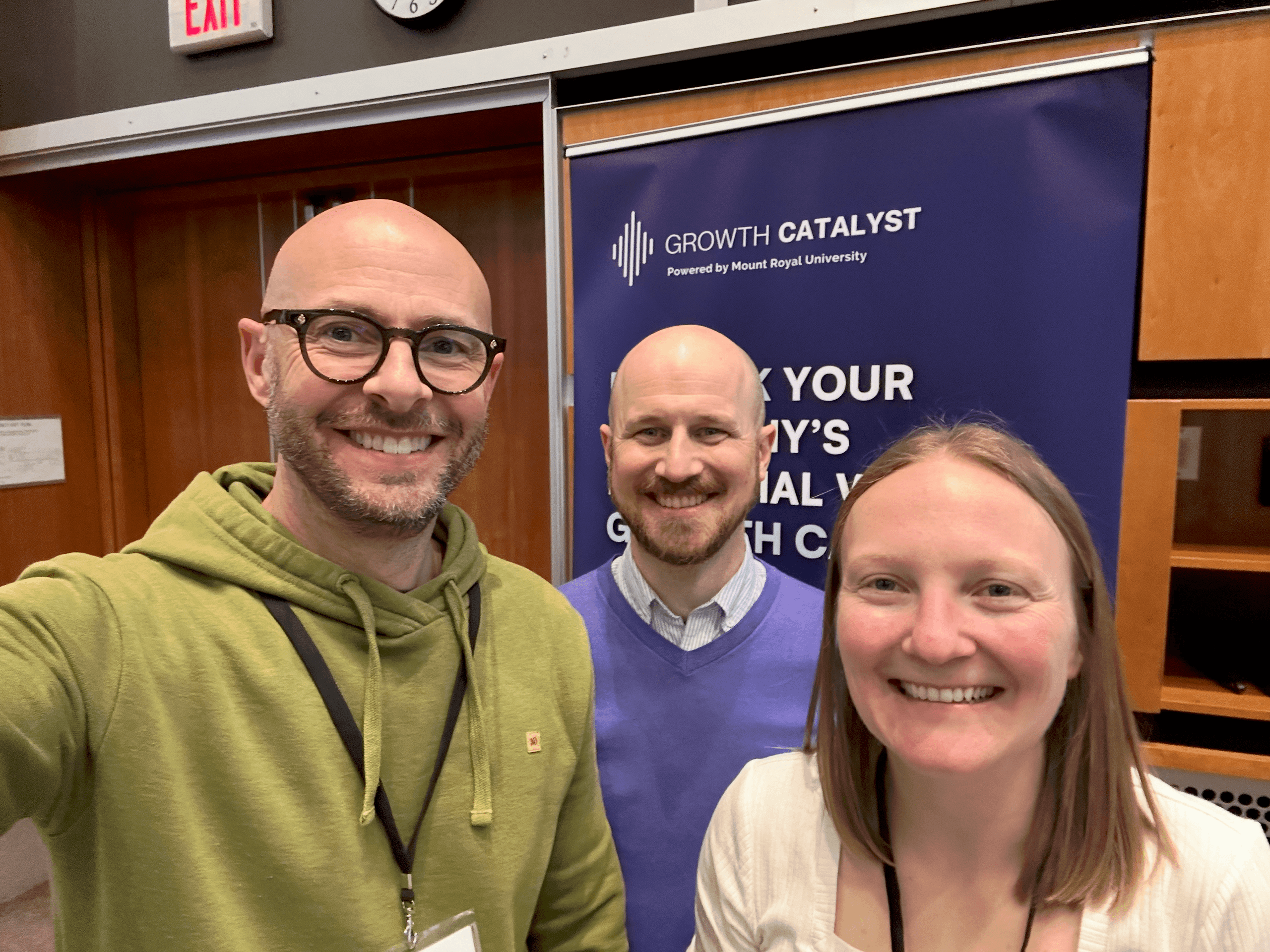 Three smiling people pose for a photo indoors at a professional event. The person on the left wears glasses and a green hoodie, the person in the middle wears a purple sweater, and the person on the right has blonde hair and is wearing a white top.