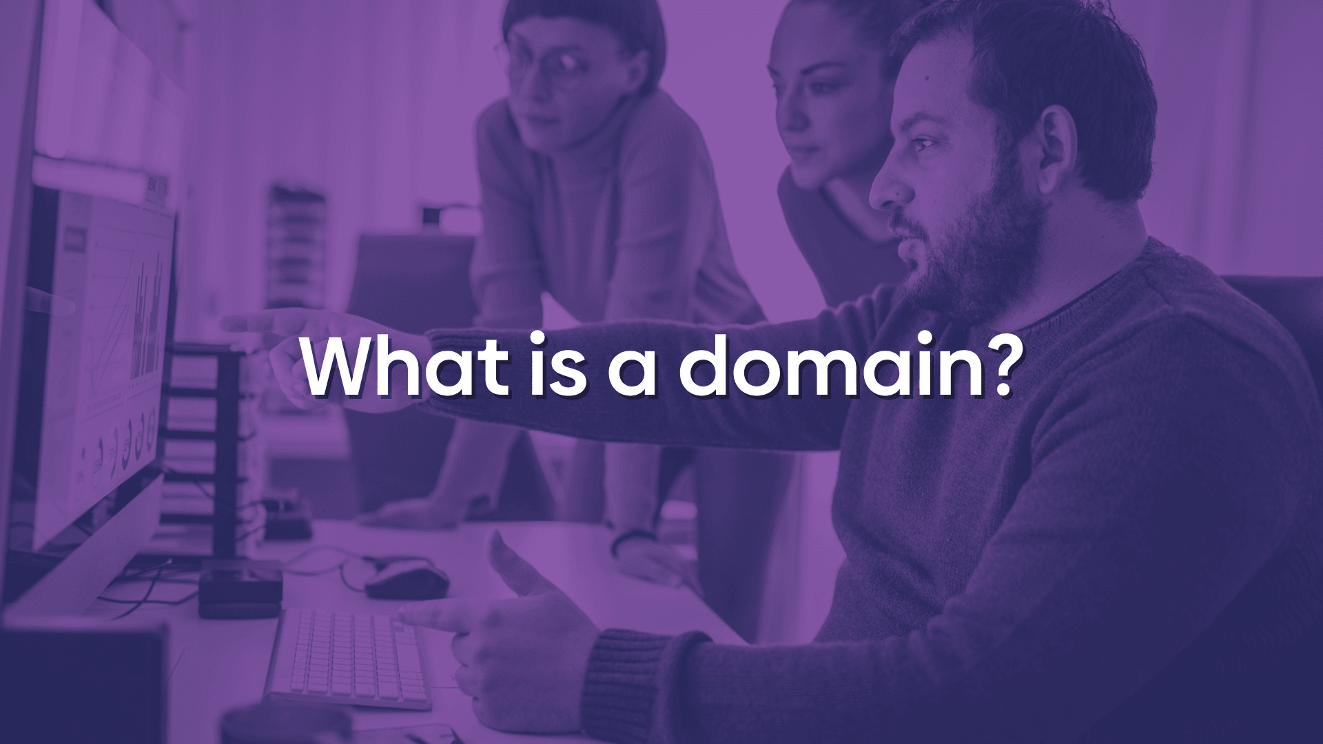 Photo of a person at a computer pointing to their screen in front of their co-workers. There is a purple overlay with text reading "What is a domain?"